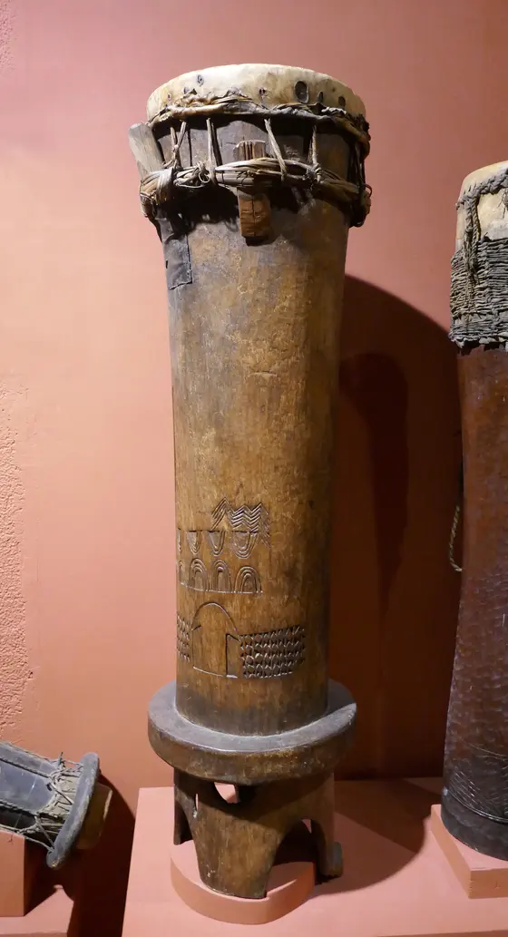 A tall drum standing on carved feet, its head lashed with cord over pegs, the body incised with bands of geometric pattern and small stylised figures.