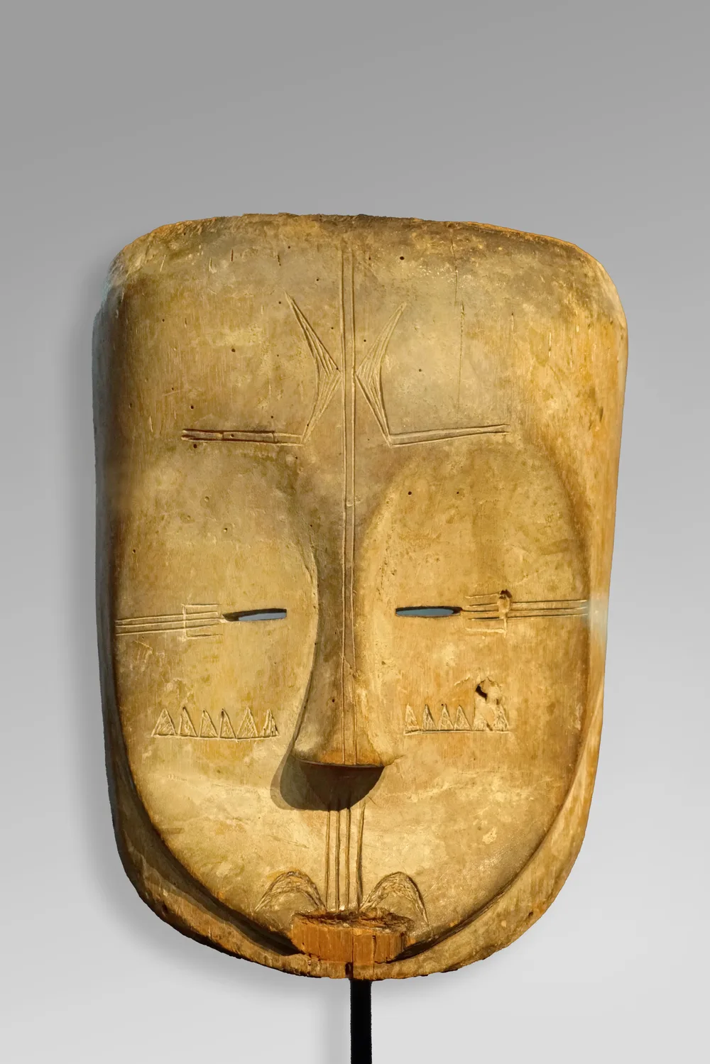 An anthropomorphic Fang mask: a long pale face coated in kaolin, the brows meeting in a continuous arc above a narrow nose, the mouth small and set low.