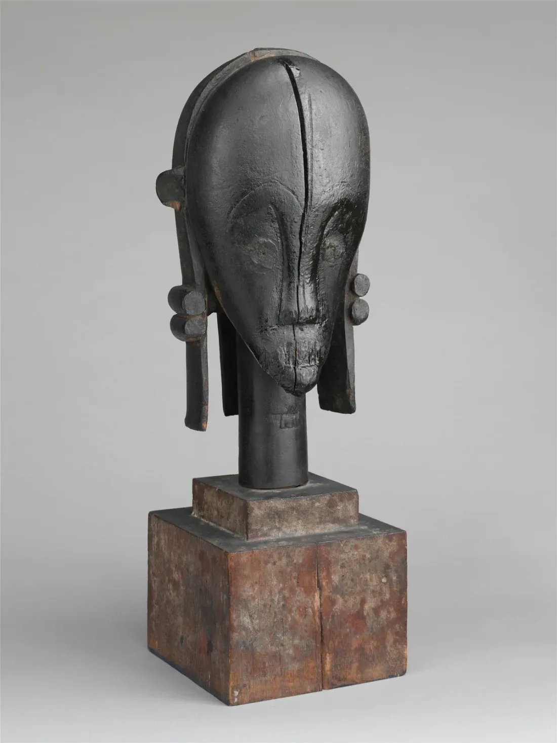 A carved dark wood head with a high domed forehead, a central crest, large projecting ears and a small pursed mouth, mounted on a worn wooden block.