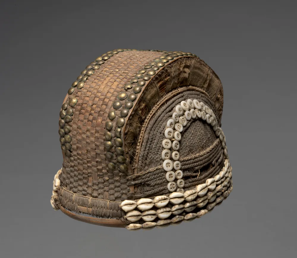 A Fang headpiece: a rounded cap built from plant fibre and cloth, its surface worked with hair, cowrie shells, buttons and upholstery studs.