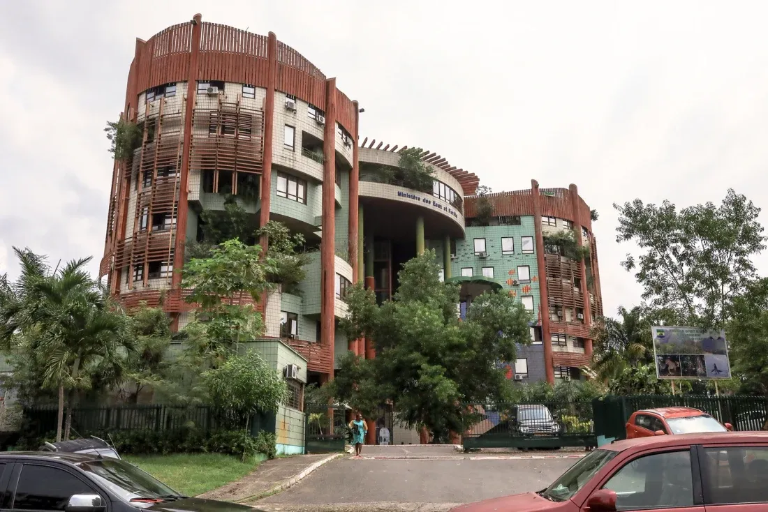 A large terracotta and green modernist office building of stacked curved balconies planted with vegetation, seen from the street behind palm trees and parked cars.