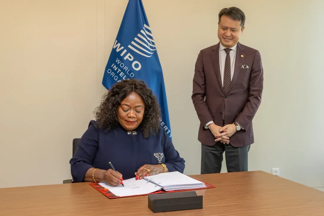 A woman in a dark suit sits at a table signing a large bound document in a red folder, while a man stands beside her watching. A World Intellectual Property Organization flag hangs behind them.