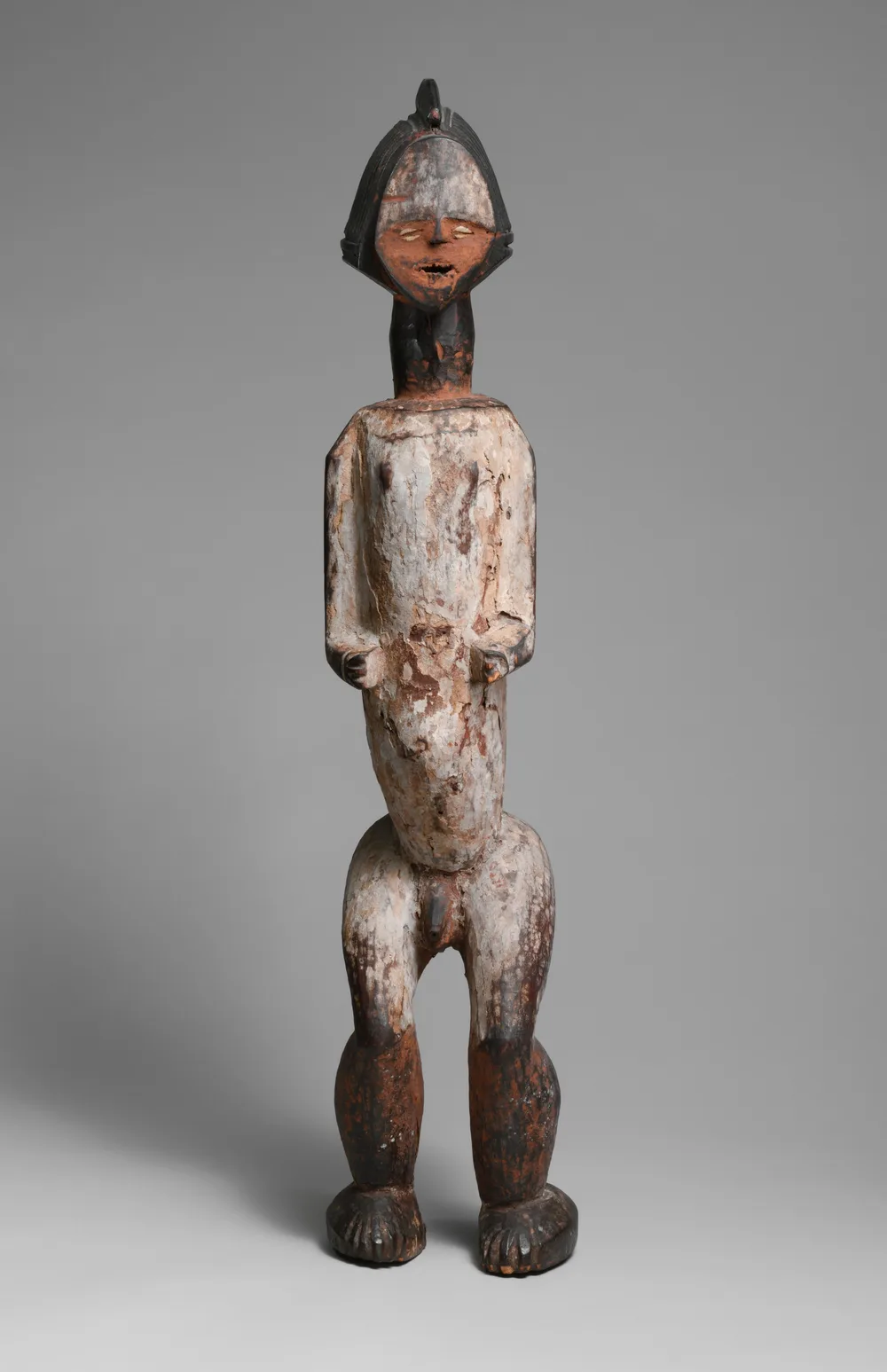 A Kota-Mbete reliquary figure: an elongated wooden body with a rounded head, surfaces darkened with pigment, set with cowrie shells and metal.