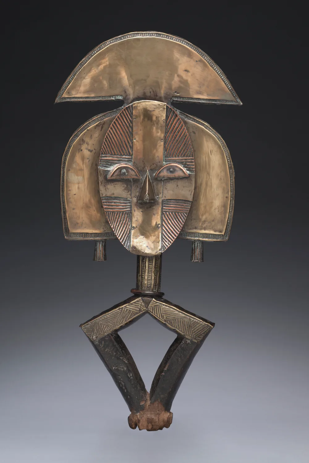 A Kota reliquary guardian figure: a flat oval face sheathed in copper alloy and iron strip, topped by a wide crescent crest, mounted on an open lozenge-shaped base.