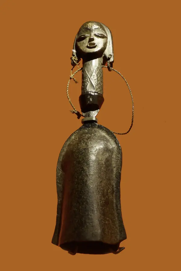 A double iron bell with two conical mouths facing opposite directions, joined by a curved handle, its surface dark and hammered.