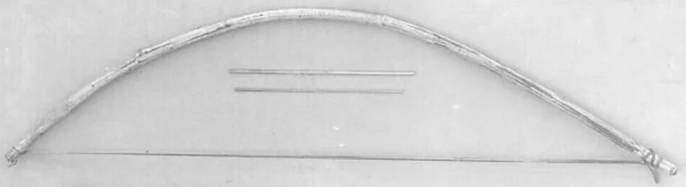 An old grey-toned photograph of a musical bow laid flat: a single curved wooden stave, bound at both ends, with a string running almost straight across its span, and two slender rods of different lengths lying beside it.