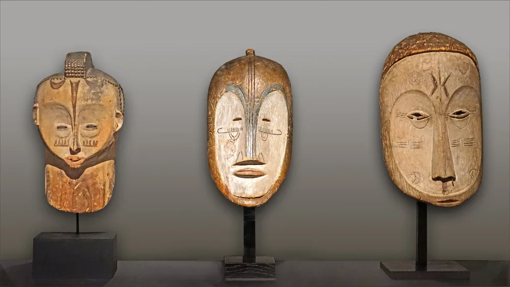 Three elongated Fang ngil masks displayed in a row behind glass: narrow white faces coated in kaolin, with long straight noses, arched brows and small closed mouths.