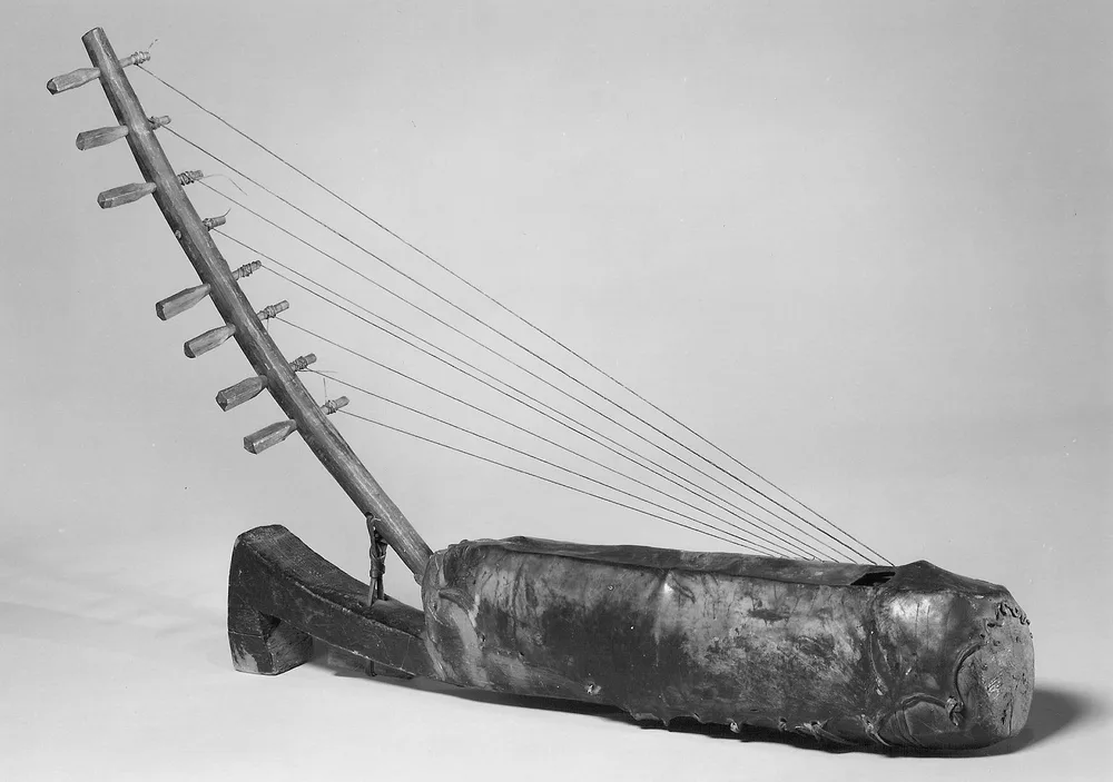 An arched harp lying on its side: a curved wooden neck rising from a boat-shaped resonator covered in stretched skin, with strings running from the neck down to the body.