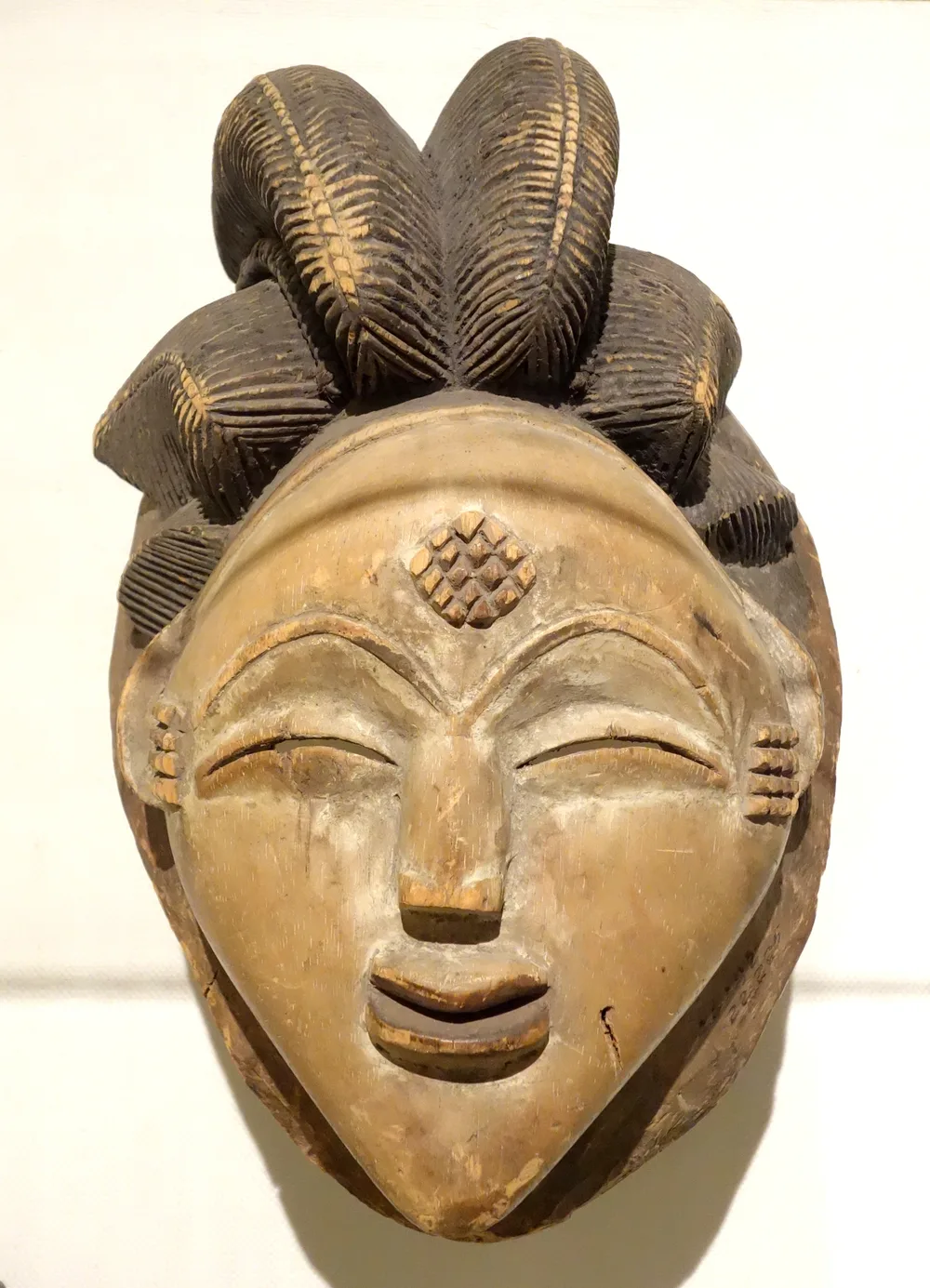 A Punu okuyi mask: a serene face whitened with kaolin, eyes narrowed to slits, lips reddened, the forehead and temples carved with raised scarification patterns, hair dressed in two high shell-shaped lobes.