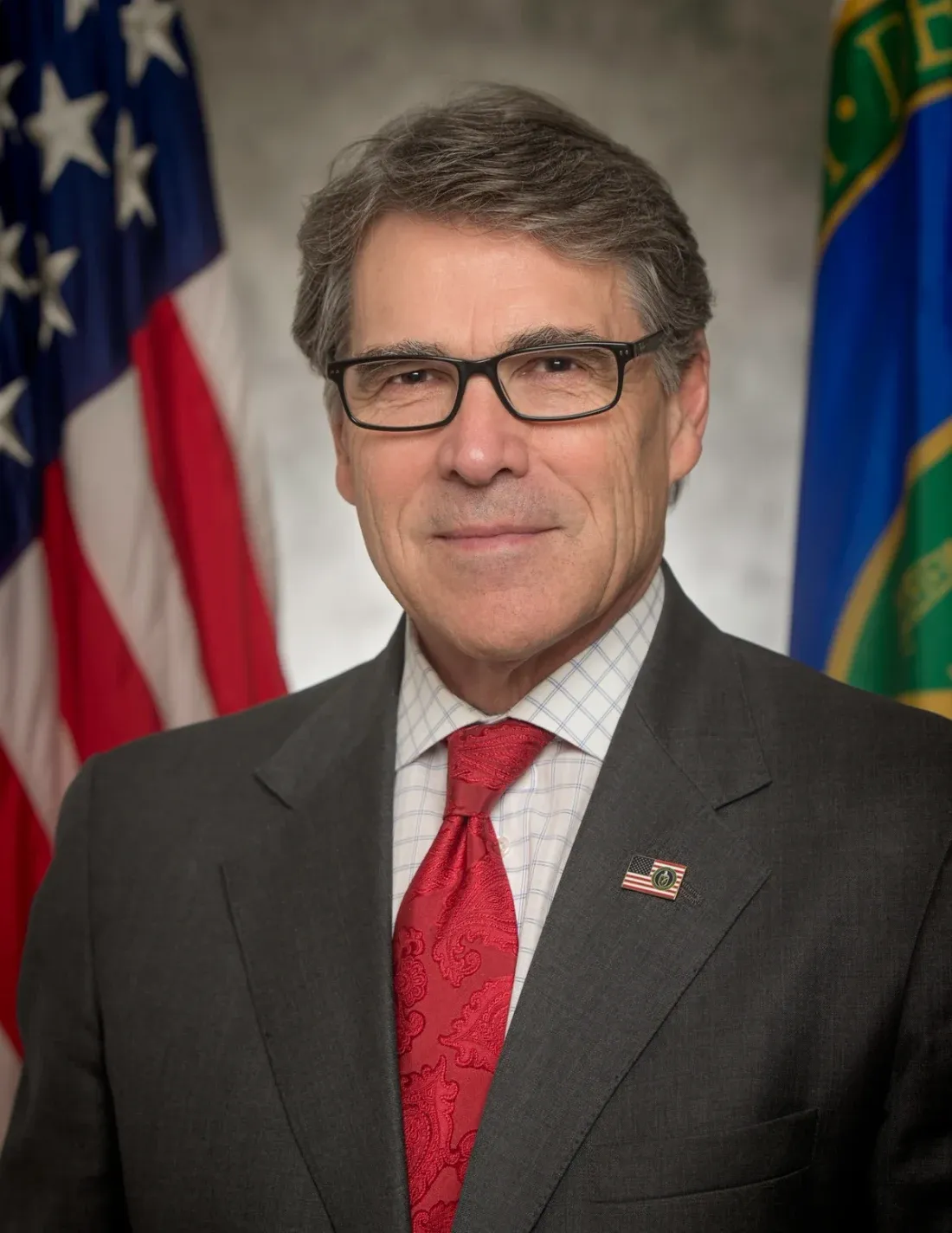 An official studio portrait of a man in a dark suit and red tie, standing in front of the United States flag and a departmental flag.