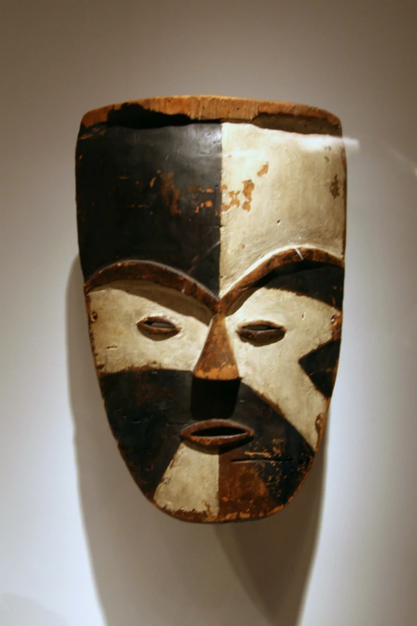 A wooden mask hanging on a plain wall, its face divided into dark and pale quarters, with narrow slit eyes, arched brows and an open oval mouth.